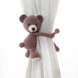 May include: A brown crocheted teddy bear with black eyes and a white nose is used as a curtain tieback. The bear is wrapped around a white sheer curtain.
