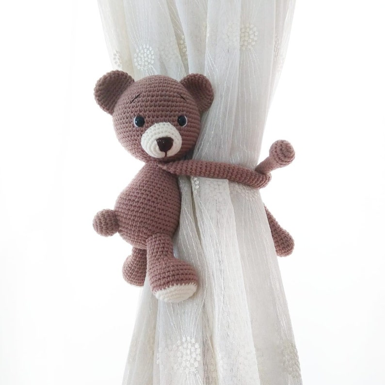 May include: A brown crocheted teddy bear with black eyes and a white nose is used as a curtain tieback. The bear is wrapped around a white sheer curtain.