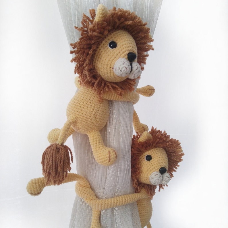 May include: Two crocheted lion curtain tiebacks in beige and brown. The larger lion is holding the smaller lion's tail.