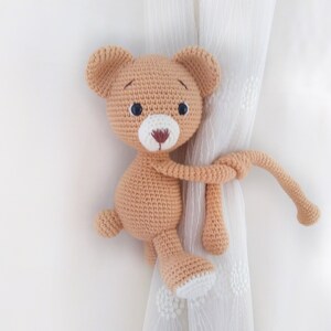 May include: A crocheted tan teddy bear with black eyes and a white nose is holding onto a white curtain with its tail.