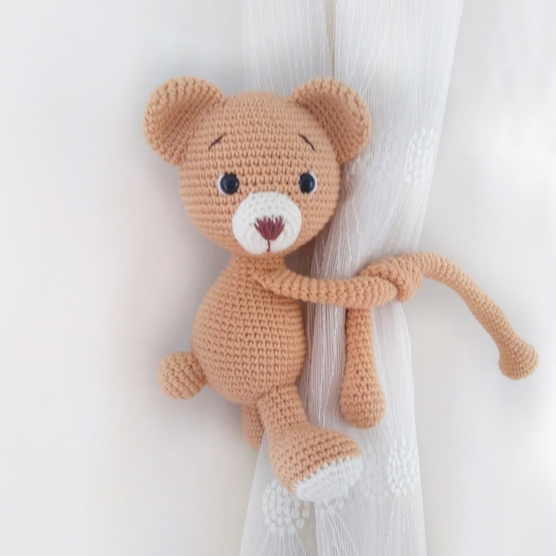 May include: A crocheted tan teddy bear with black eyes and a white nose is holding onto a white curtain with its tail.