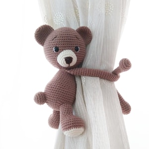 May include: A brown crocheted teddy bear with black eyes and a white nose is used as a curtain tieback. The bear is holding onto a white sheer curtain.