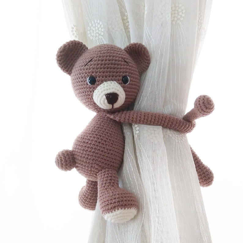 May include: A brown crocheted teddy bear with black eyes and a white nose is used as a curtain tieback. The bear is holding onto a white sheer curtain.