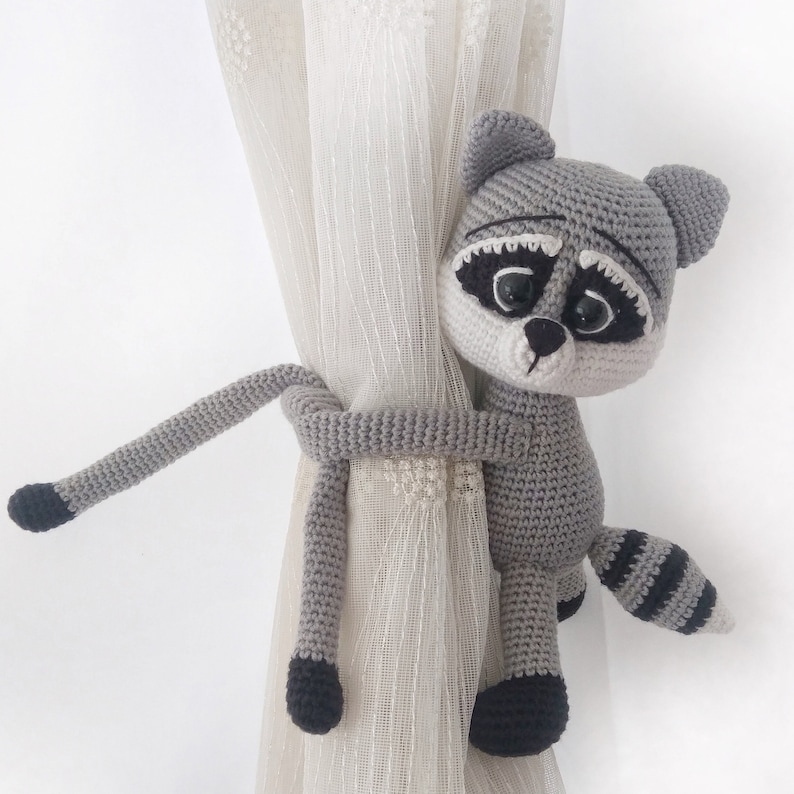May include: A crocheted raccoon curtain tieback in gray and black. The raccoon is holding onto a sheer white curtain with its tail wrapped around the curtain rod.