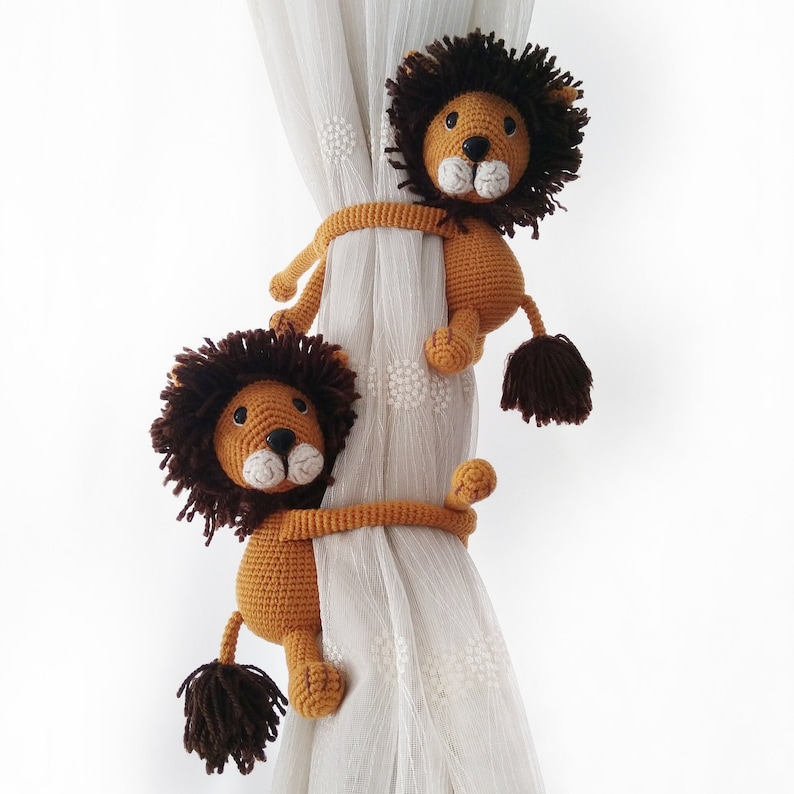 May include: Two crocheted lion curtain tiebacks in brown and white. The lions are holding onto a white curtain with their paws.