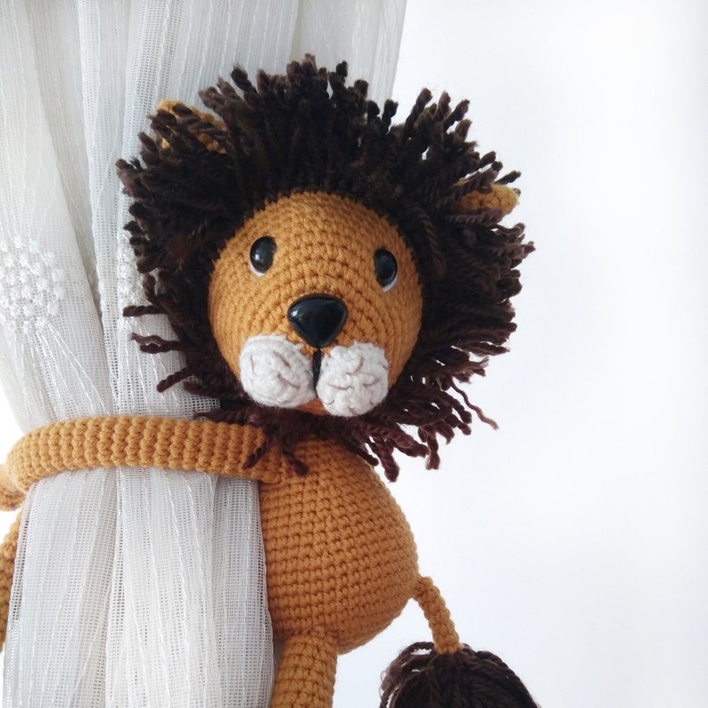 May include: A crocheted lion toy with brown fur and a tan body. The lion is holding onto a white curtain with its paws.