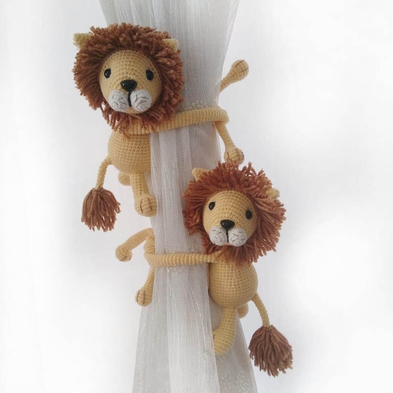 May include: Two crocheted lion curtain tiebacks in beige and brown with white faces. The tiebacks are holding up a sheer white curtain.