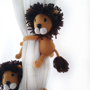 May include: A brown crocheted lion toy with a long tail and a fluffy mane. The lion is holding onto a white curtain with its paws.