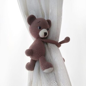May include: A brown crocheted teddy bear with a white nose and black eyes is used as a curtain tieback. The bear is holding onto a white sheer curtain.