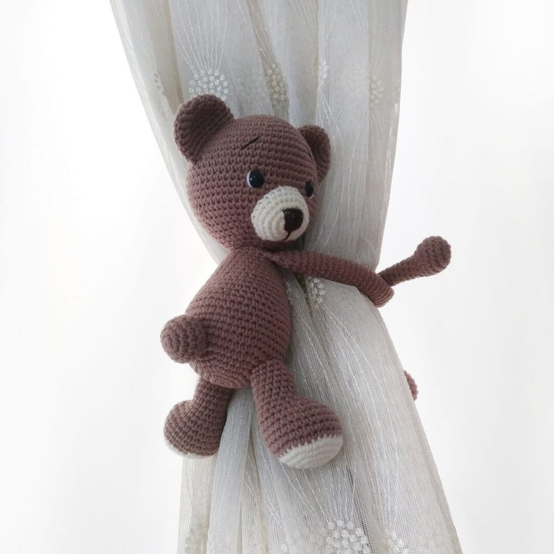 May include: A brown crocheted teddy bear with a white nose and black eyes is used as a curtain tieback. The bear is holding onto a white sheer curtain.