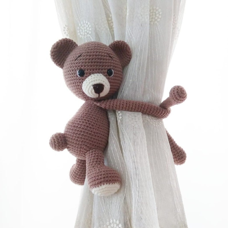 May include: A brown crocheted teddy bear with white accents is used as a curtain tieback. The bear is holding onto a white sheer curtain with its arms and legs.