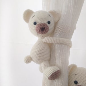 May include: A white crocheted teddy bear with brown eyes and a brown nose is holding onto a white curtain. The bear is used as a curtain tieback.
