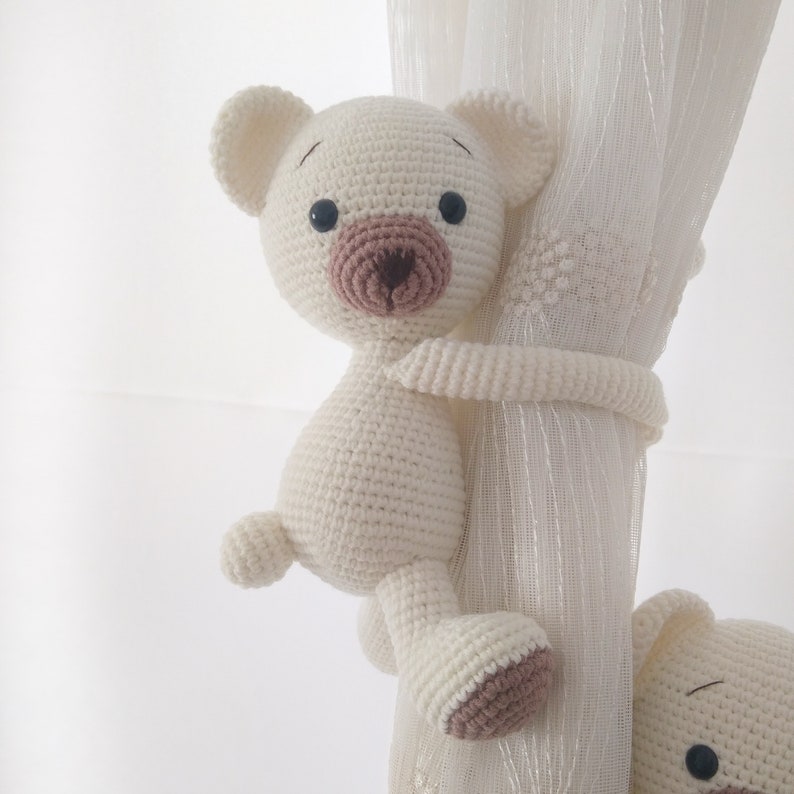 May include: A white crocheted teddy bear with brown eyes and a brown nose is holding onto a white curtain. The bear is used as a curtain tieback.