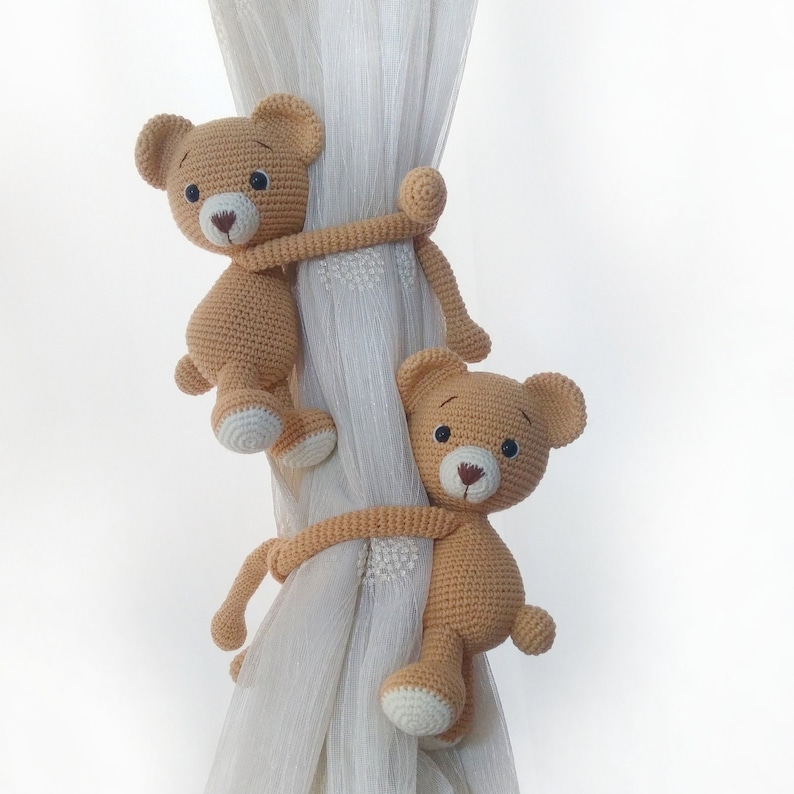 May include: Two crocheted teddy bears in beige are hanging from a white sheer curtain. The bears are holding onto the curtain with their paws and are facing each other.