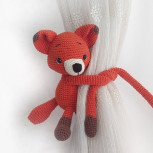 May include: A crocheted orange fox toy with brown accents is holding onto a white curtain. The fox's tail is wrapped around the curtain and the toy is used as a curtain tieback.