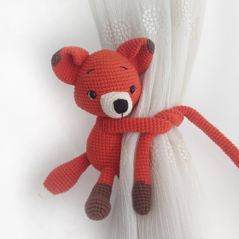 May include: A crocheted orange fox toy with brown accents is holding onto a white curtain. The fox's tail is wrapped around the curtain and the toy is used as a curtain tieback.
