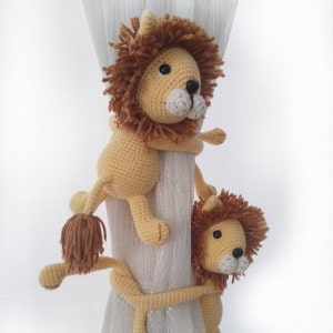 May include: Two crocheted lion curtain tie backs in beige and brown. The lions are holding onto a white curtain rod.