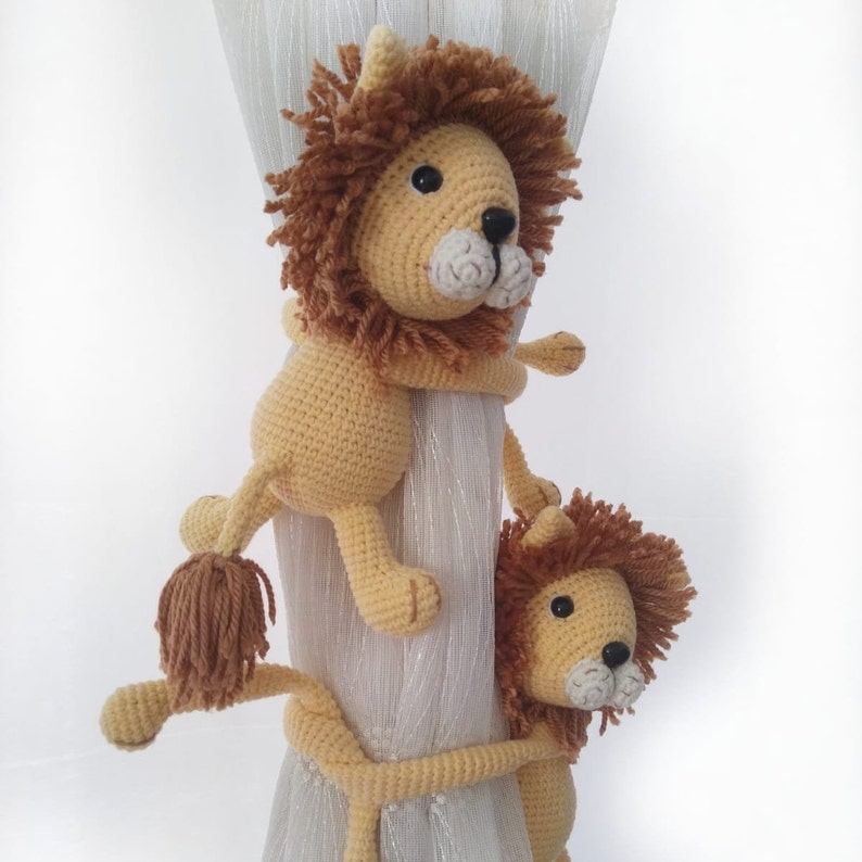 May include: Two crocheted lion curtain tie backs in beige and brown. The lions are holding onto a white curtain rod.