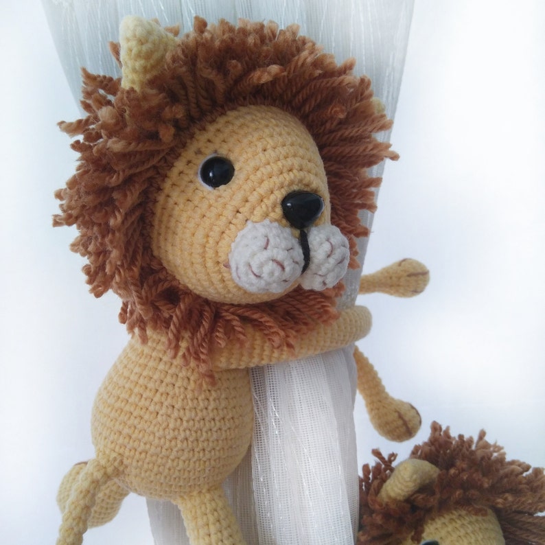 May include: A crocheted lion toy with a brown mane and a white face. The lion is holding onto a white curtain with its paws.