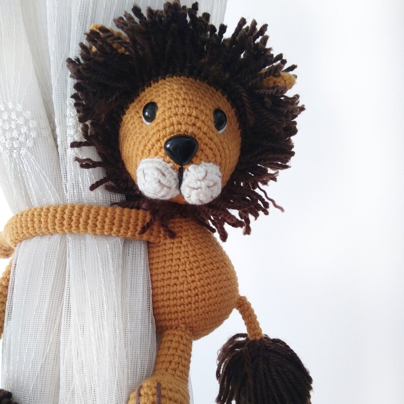 May include: A crocheted lion toy with brown fur and a tan body. The lion is holding onto a white curtain with its paws.