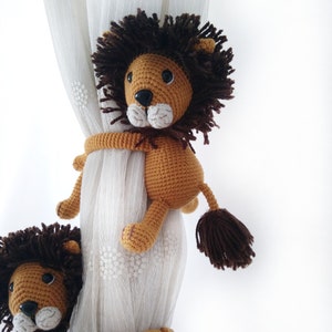 May include: A brown crocheted lion toy with a long tail and a fluffy mane is holding onto a white sheer curtain. The lion has black eyes and a white nose.