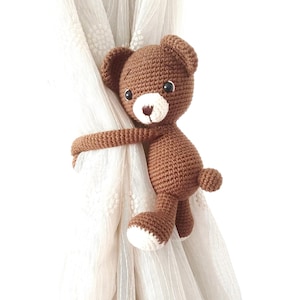 May include: A brown crocheted teddy bear curtain tieback. The bear is holding onto a white sheer curtain with its arms.