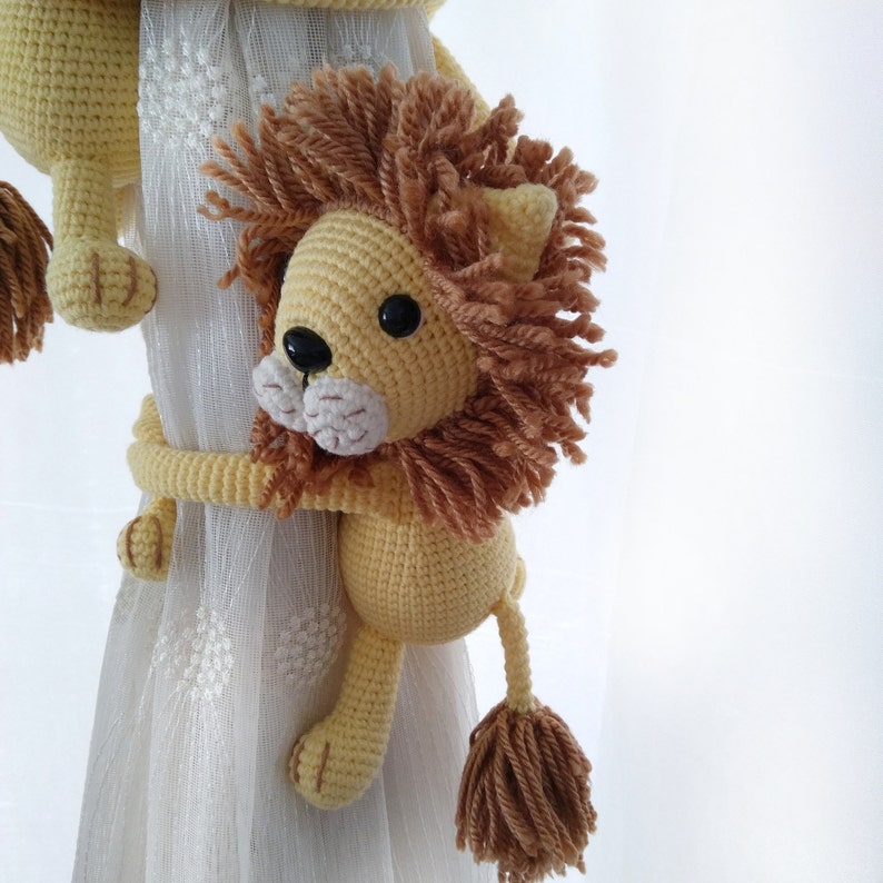 May include: A crocheted lion toy with a brown mane and tail, holding onto a white curtain. The lion is yellow with black eyes and a white nose.