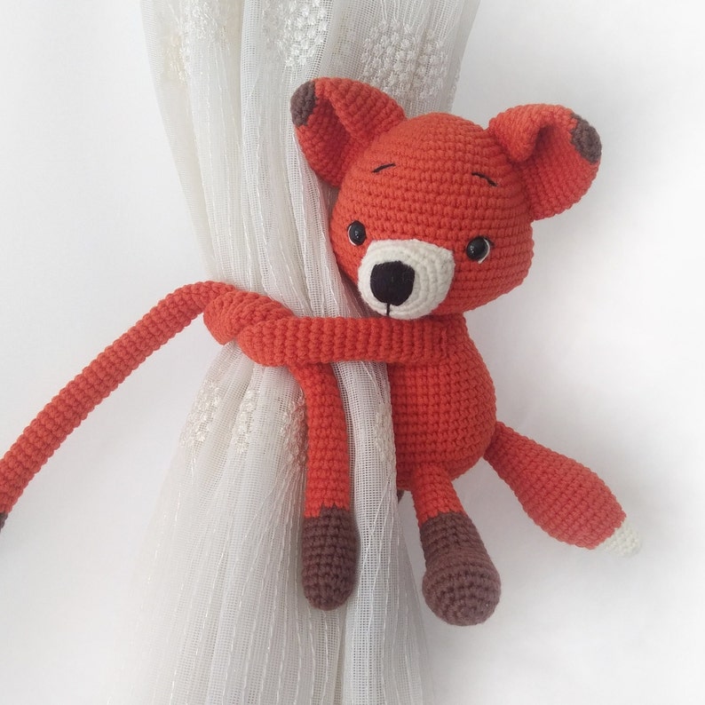 May include: Orange crocheted fox toy with brown accents, holding a white curtain with its paws. The fox is used as a curtain tieback.