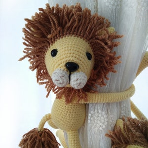 May include: A crocheted lion toy with a yellow body and brown mane. The lion is holding onto a white crocheted curtain with its paws.