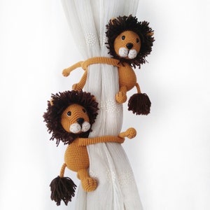 May include: Two crocheted lion curtain tiebacks in brown and tan with brown eyes and black noses. The lions are holding onto a white curtain.