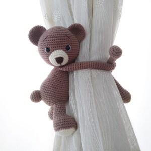 May include: A brown crocheted teddy bear with black eyes and a white nose is used as a curtain tieback. The bear is holding onto a white sheer curtain with its arms.