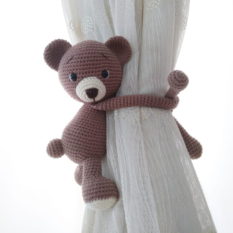 May include: A brown crocheted teddy bear with black eyes and a white nose is used as a curtain tieback. The bear is holding onto a white sheer curtain with its arms.