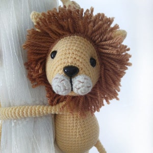 May include: A crocheted lion toy with a brown mane and a light brown body. The lion is holding onto a white curtain.
