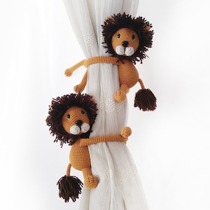 May include: Two crocheted lion curtain tiebacks in brown and tan. The lions are holding onto a white curtain with their paws.