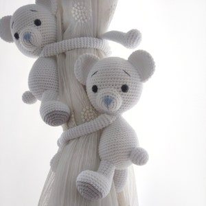 May include: Two white crocheted teddy bears with blue noses are used as curtain tiebacks. The bears are holding onto a white sheer curtain.