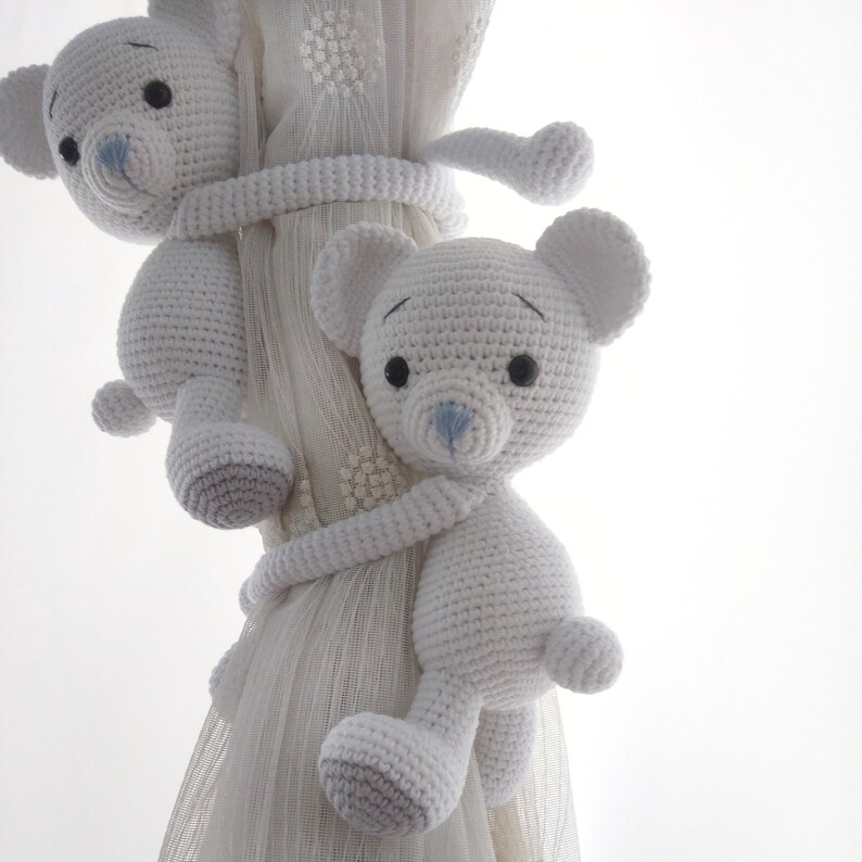 May include: Two white crocheted teddy bears with blue noses are used as curtain tiebacks. The bears are holding onto a white sheer curtain.