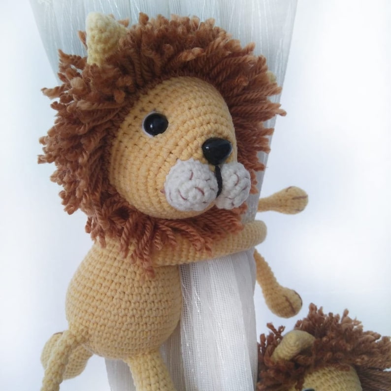 May include: A crocheted lion toy with a brown mane and a beige body. The lion is holding onto a white curtain with its paws.