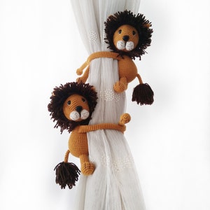 May include: Two brown crocheted lion curtain tie backs with brown manes and white faces. The lions are holding onto a white sheer curtain.