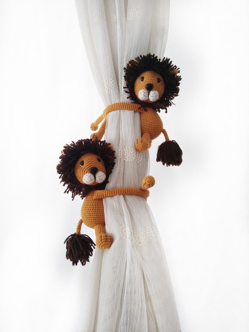 May include: Two brown crocheted lion curtain tie backs with brown manes and white faces. The lions are holding onto a white sheer curtain.