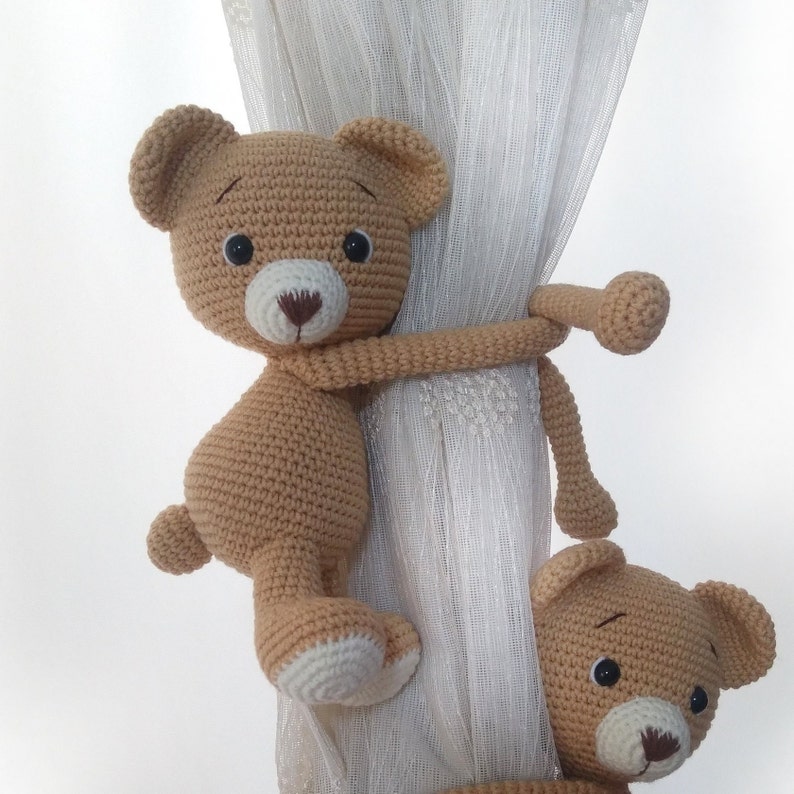 May include: Two crocheted teddy bears in brown, one hanging from a white curtain with a brown crocheted tail.