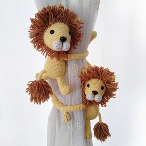 May include: Two crocheted lion curtain tie backs in yellow and brown. The lions are holding onto a white curtain with a floral pattern.