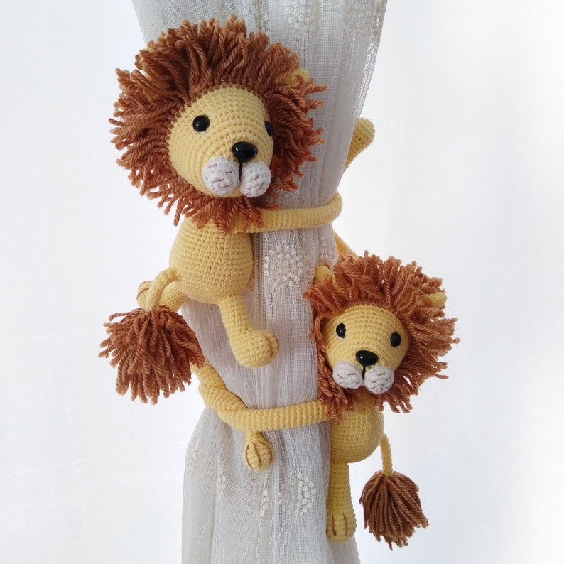 May include: Two crocheted lion curtain tie backs in yellow and brown. The lions are holding onto a white curtain with a floral pattern.