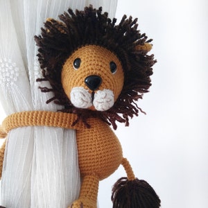 May include: A crocheted lion toy with brown fur and a tan body, holding onto a white curtain with its arms. The lion has black eyes and a white nose.
