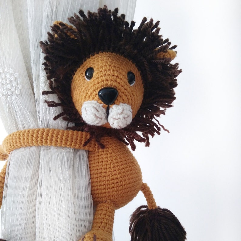 May include: A crocheted lion toy with brown fur and a tan body, holding onto a white curtain with its arms. The lion has black eyes and a white nose.