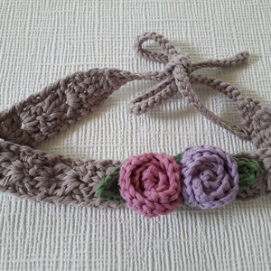 May include: A crocheted headband with a brown base and a tie closure. The headband is decorated with three crocheted flowers: two in shades of pink and purple, and one green.