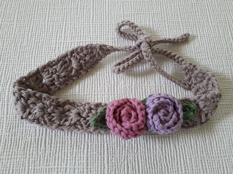 May include: A crocheted headband with a brown base and a tie closure. The headband is decorated with three crocheted flowers: two in shades of pink and purple, and one green.