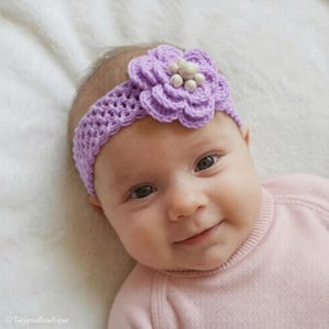 May include: A baby wearing a lavender crochet headband with a large flower and small bead accents. The headband is on the baby's head, and the baby is wearing a pink sweater. The baby is smiling.