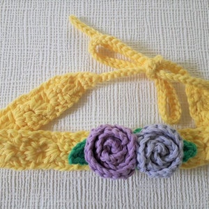 May include: A yellow crocheted headband with two purple crocheted flowers and green leaves.