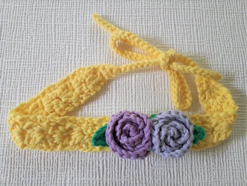 May include: A yellow crocheted headband with two purple crocheted flowers and green leaves.