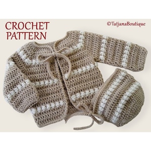 May include: A crocheted baby cardigan and hat set in beige and white. The cardigan has a tie closure and the hat is a simple beanie style.
